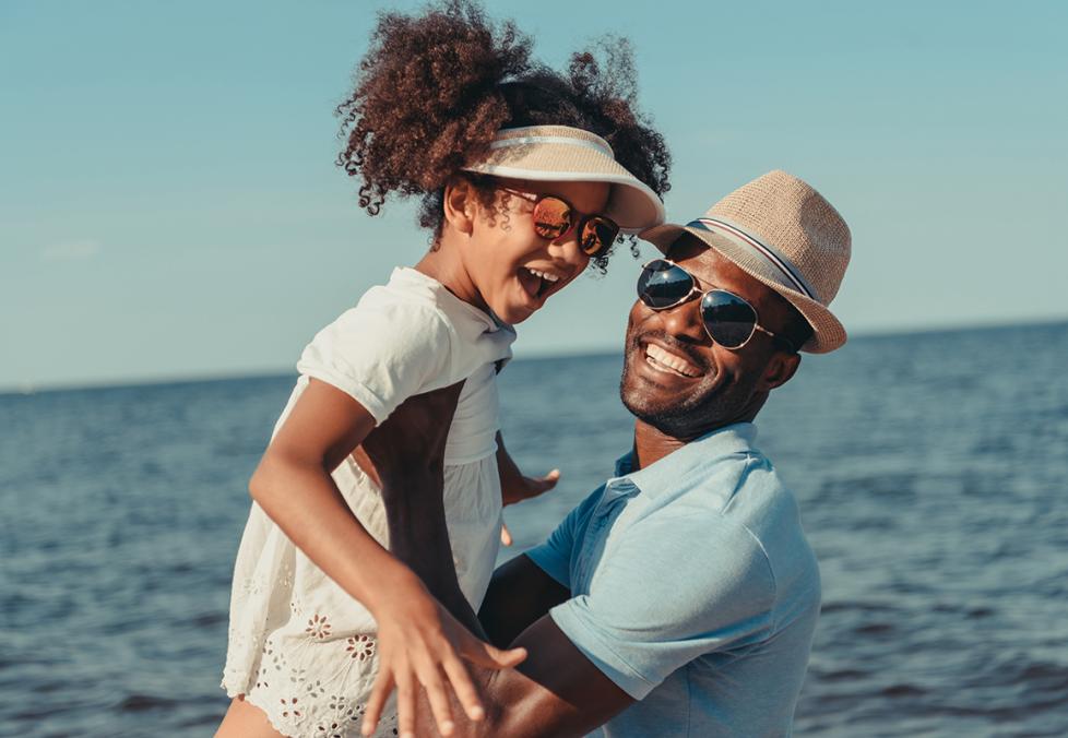 A father holding up his daughter at the beach, both wearing sunglasses